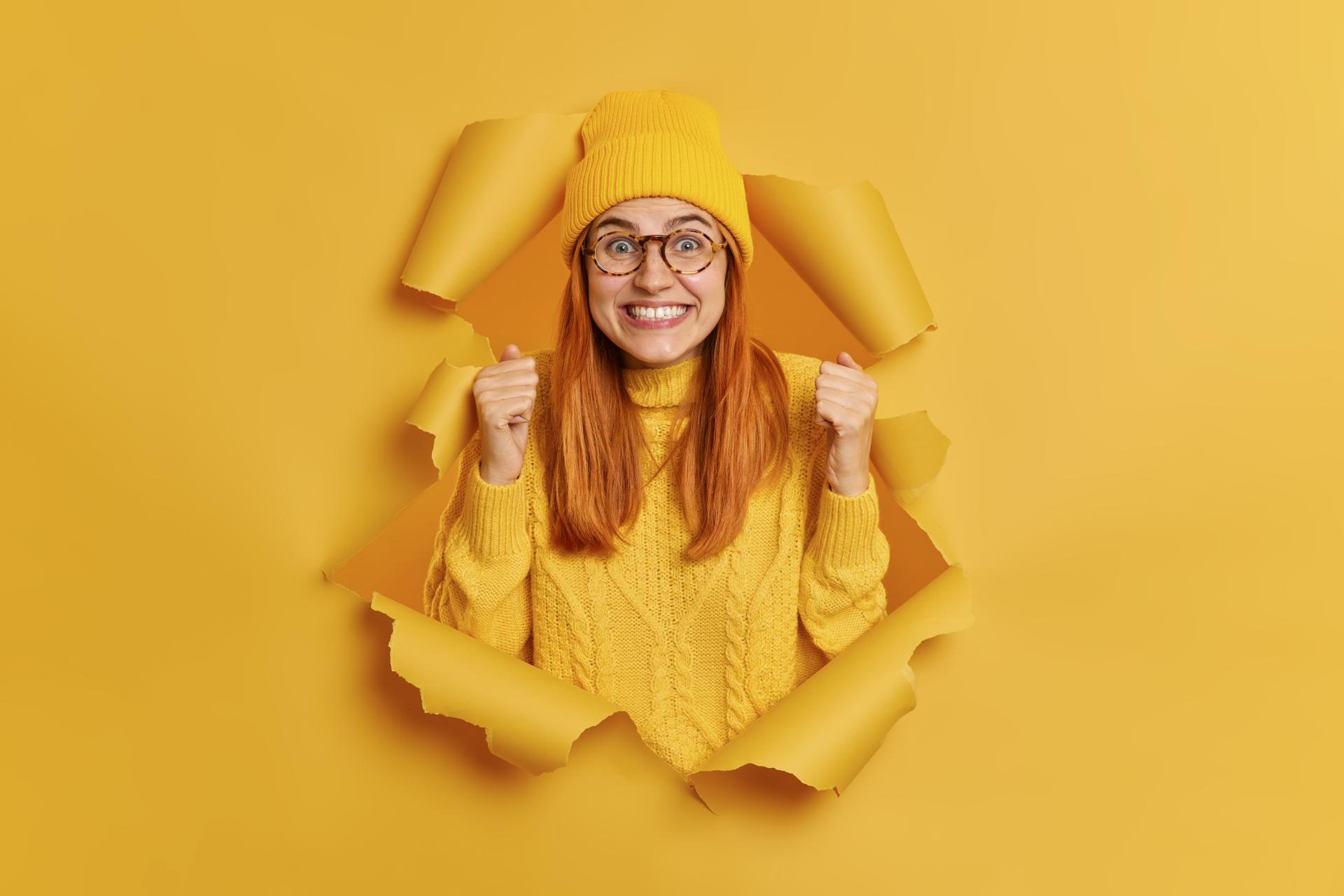 Positive excited redhead woman raises fists wears yellow hat knitted jumper expresses joy breaks through paper wall edited