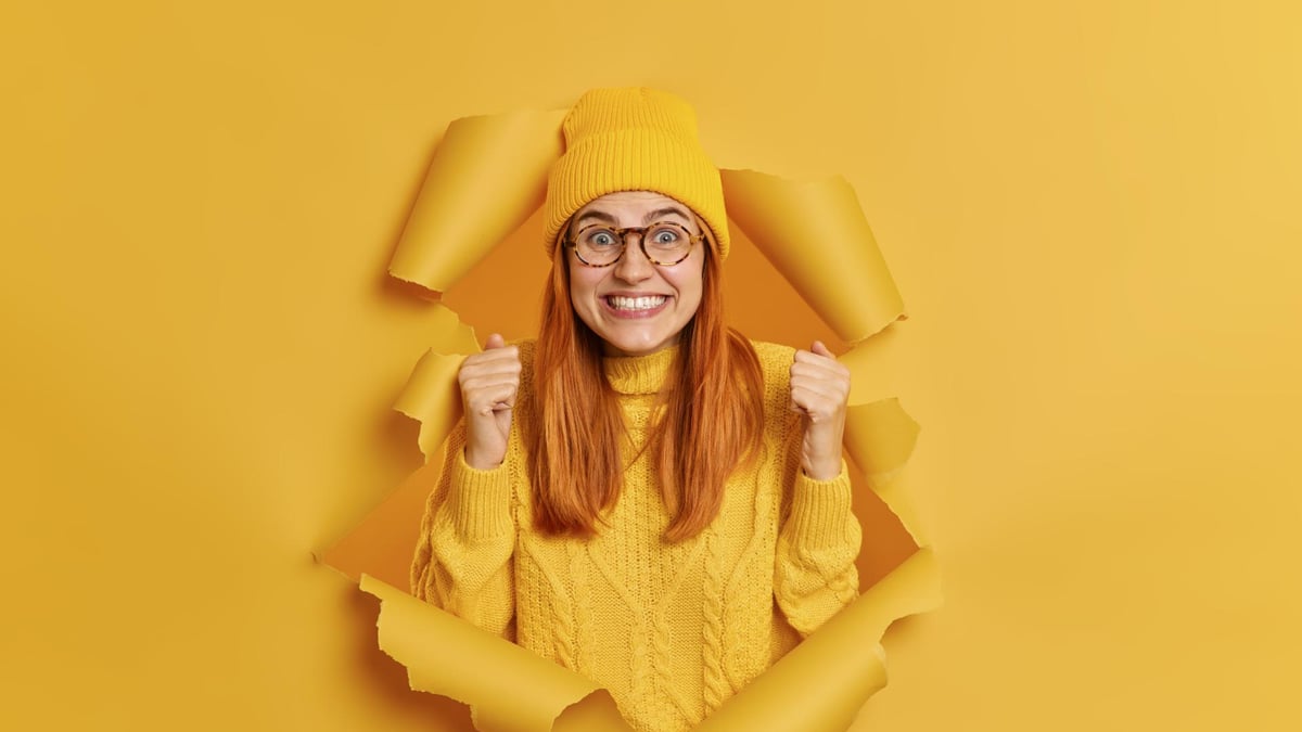 Positive excited redhead woman raises fists wears yellow hat knitted jumper expresses joy breaks through paper wall edited
